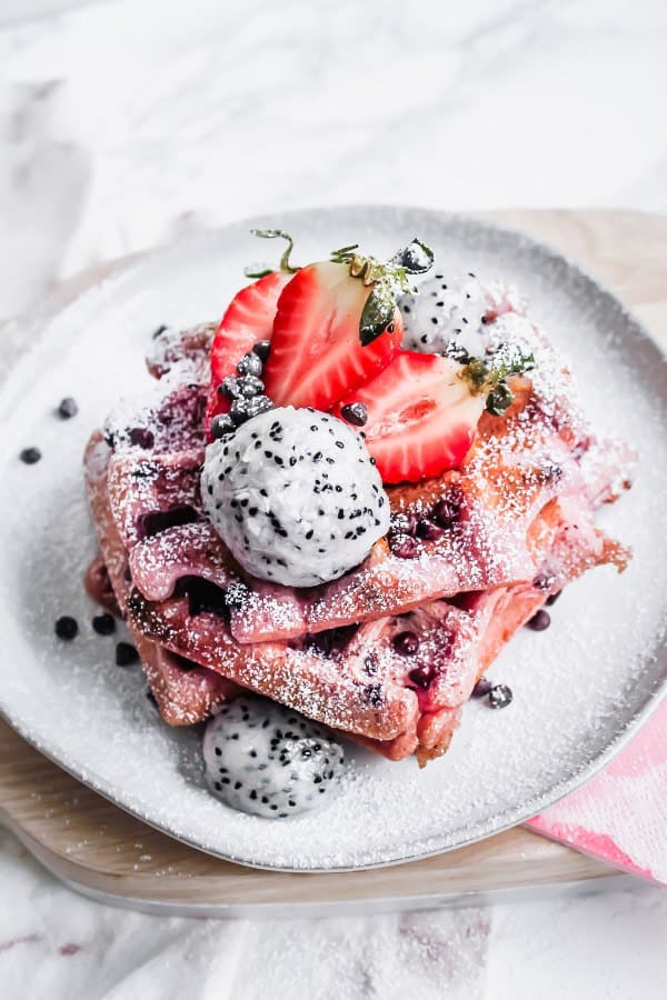 pink dragonfruit chocolate chip waffles on a plate with three strawberries, a ball of dragon fruit and sprinkled powdered sugar