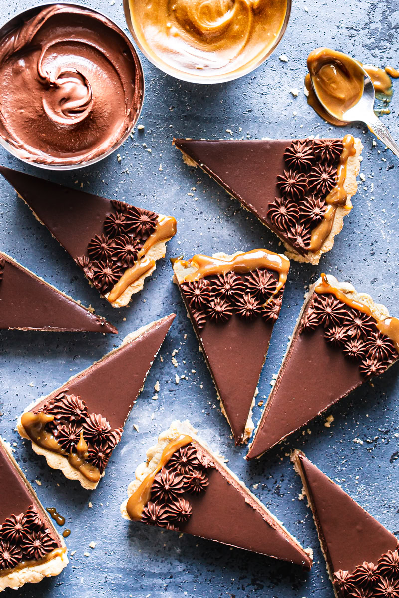 slices of Twix tart piped with chocolate mousse on a blue backdrop with a bowl of chocolate mousse and a bowl of vegan caramel