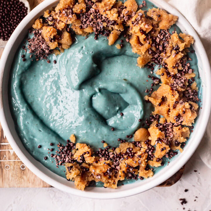a blue spirulina smoothie bowl topped with almond flour cookie dough, chocolate covered chia seeds on top of a wooden cutting board with a bowl of cookie dough and cacao nibs beside it