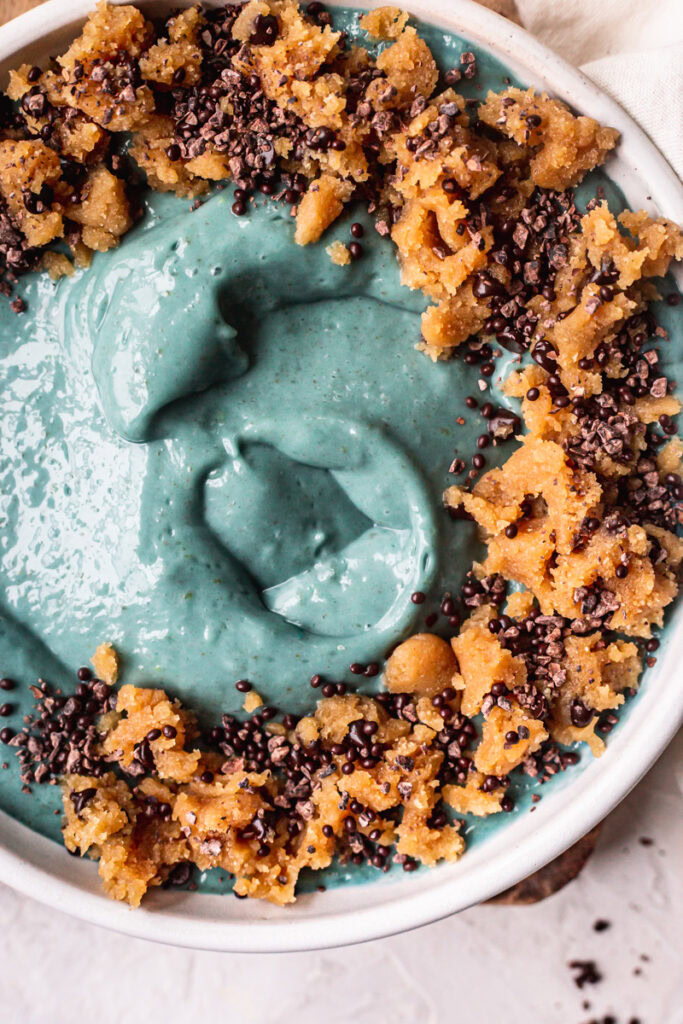 a close up of a blue spirulina smoothie bowl topped with almond flour cookie dough, chocolate covered chia seeds