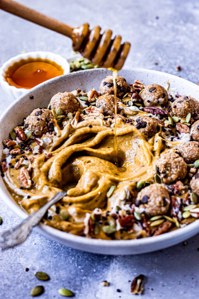 an action shot of a pumpkin pie smoothie in a bowl topped with cookie dough balls, pumpkin seeds, pecans, chocolate covered chia seeds being drizzled with maple syrup
