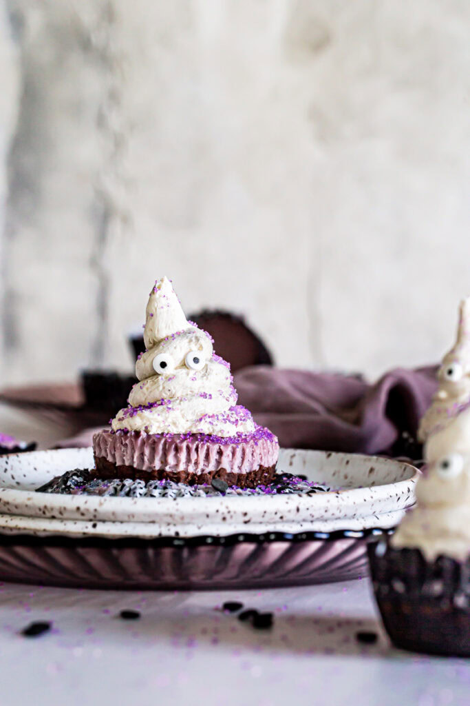 cheesecake ghost on a plate in the middle of the frame with 3 other cheesecakes out of focus in front and behind with a purple linen towel in the background