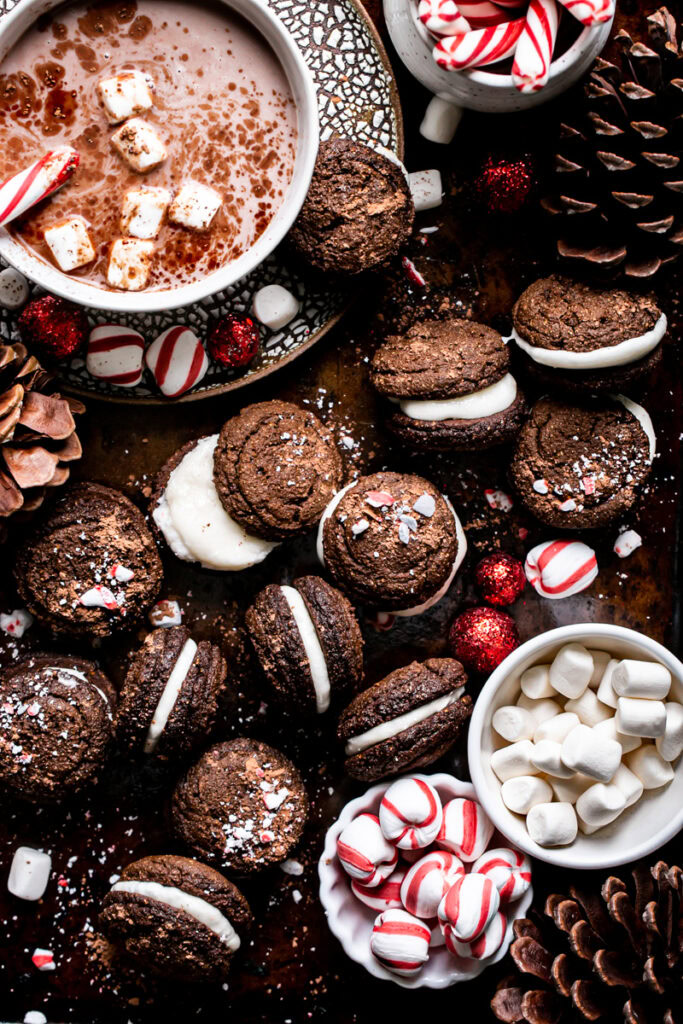 a tray of hot cocoa whoopie pies with a mug of hot cocoa in the upper left and a bowl of peppermint candies and marshmallows at the bottom right