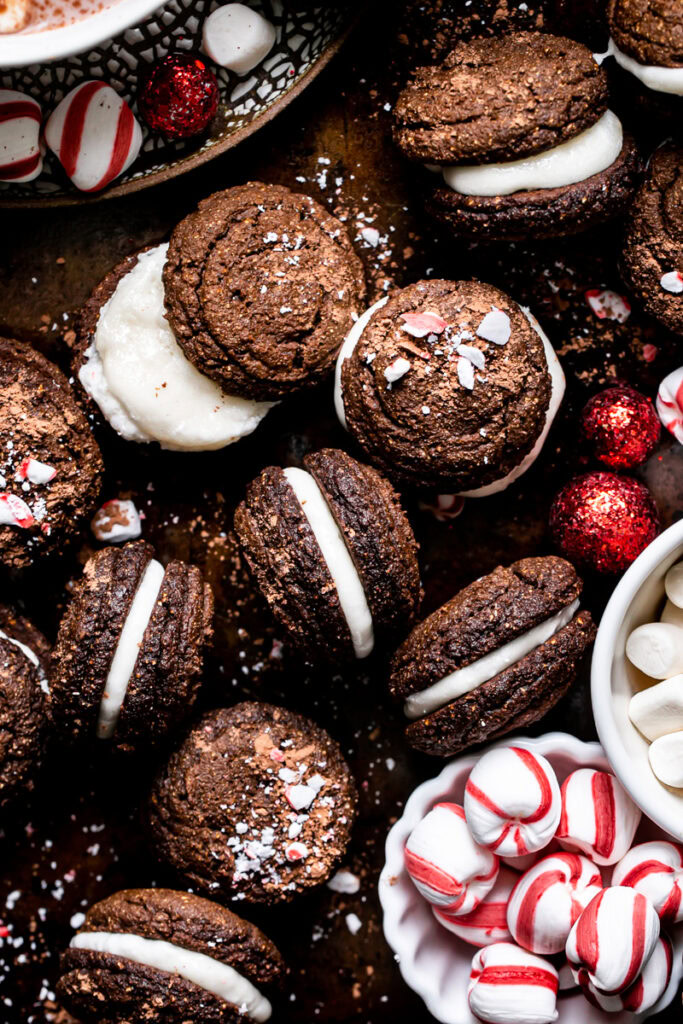 hot cocoa whoopie pies piled on a baking sheet with and peppermint candies and vegan marshmallows in the bottom right