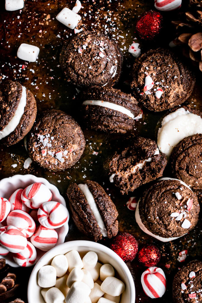 hot cocoa whoopie pies piled on a baking sheet, one with a bit taken out of it. A bowl of peppermint candies and vegan marshmallows in the bottom left