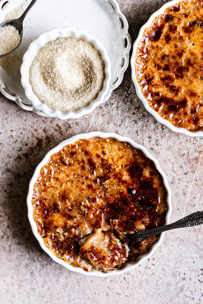 two dishes of vegan gingerbread creme brûlée with a bowl of sugar on a plate next to it and a spoon scooping some custard out