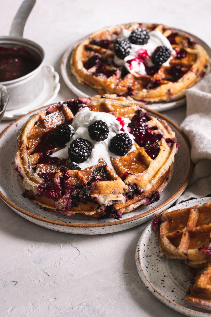 3/4 view of 3 plates of blackberry waffles with a saucepan of blackberry syrup in the background