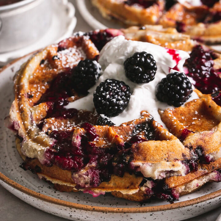close up side view of a plate of blackberry waffles with more in the background and a pot of blackberry syrup