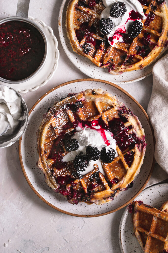 overhead image of 3 plates of blackberry waffles with a bowl of whipped cream and a pot of homemade blackberry syrup next to it