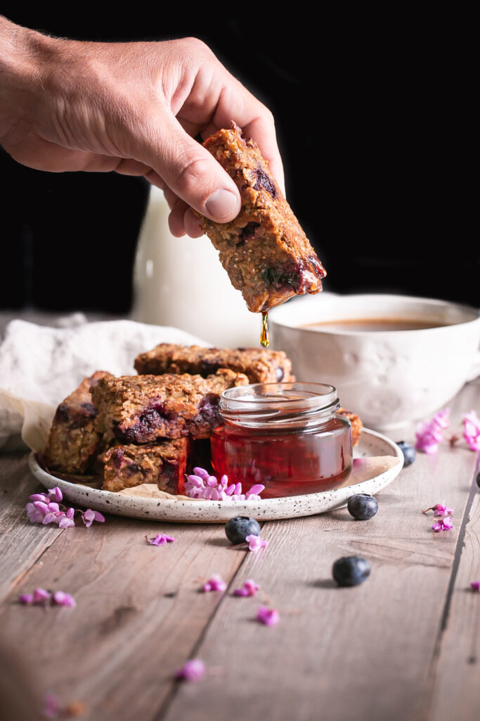 Blueberry Oatmeal French Toast Sticks on a plate one in hand being dipped in a jar of maple syrup with a jar of maple syrup beside it and a cup of coffee and jug of almond milk in the background