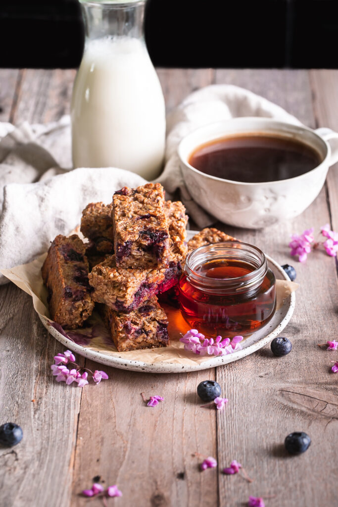 3/4 image of Blueberry Oatmeal French Toast Sticks on a plate with a jar of maple syrup beside it and a cup of coffee and jug of almond milk in the background 