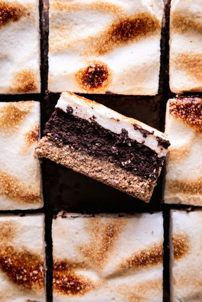 sliced s'mores brownies with toasted marshmallow fluff on top and the middle brownie on its side showing the brownie center and graham-style crust