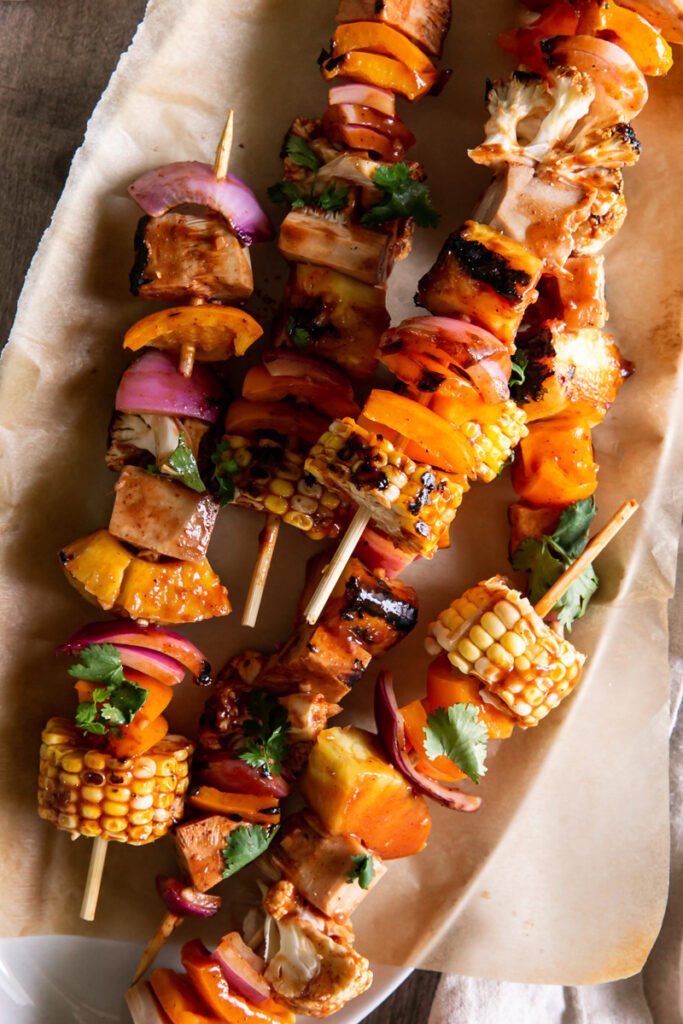 close up of an oval platter with grilled veggie barbecue kabobs on it with a side of barbecue sauce in a bowl beside it and fresh cilantro on a plate above it
