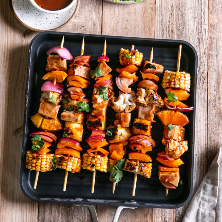 Grilled veggie barbecue jackfruit kabobs in a square Calphalon grilling pan with a bowl of barbecue sauce beside it and a plate of cilantro above it