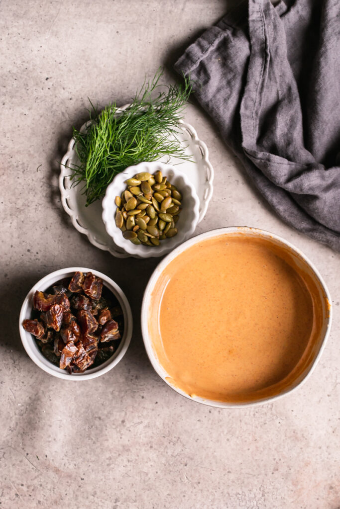 ingredients used to make the recipe. The tahini harissa sauce in a bowl, a bowl of chopped dates, a bowl of toasted pumpkin seeds, and fresh dill