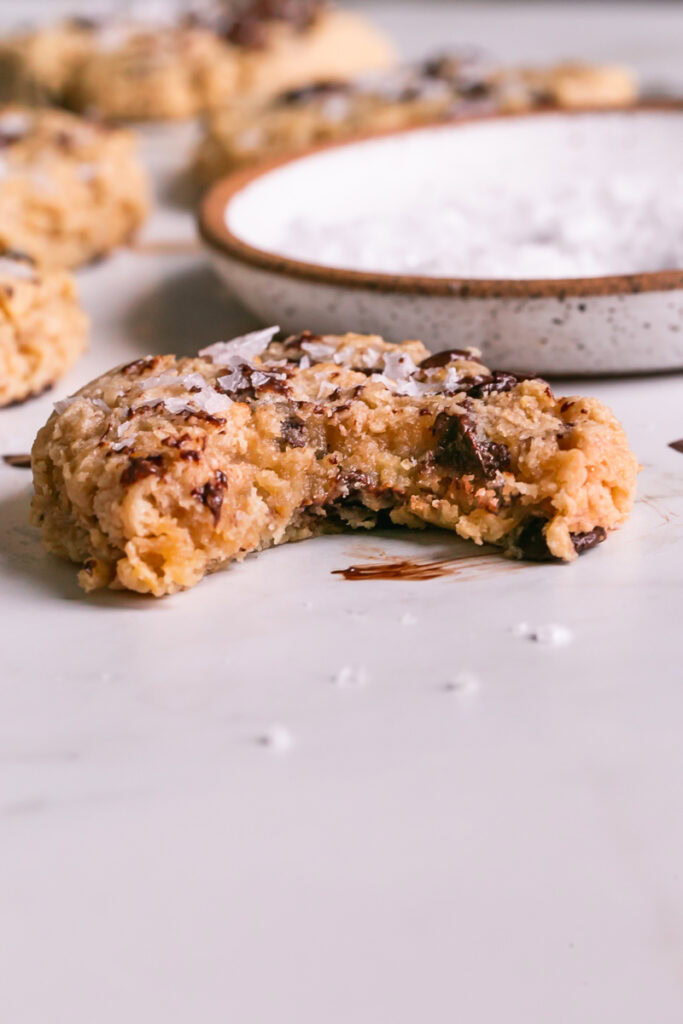 a single flourless salted tahini chocolate chip cookie with a bite taken out of it and other cookies in the background with a bowl of salt