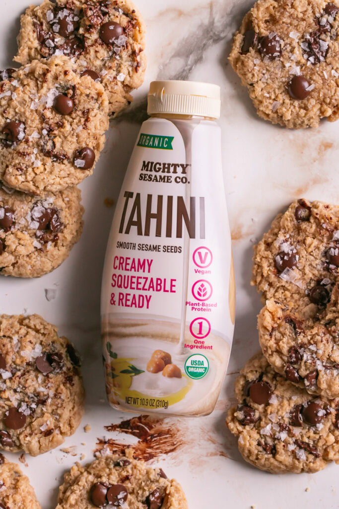 a bottle of mighty sesame co organic tahini with flourless salted tahini chocolate chip cookies all around it