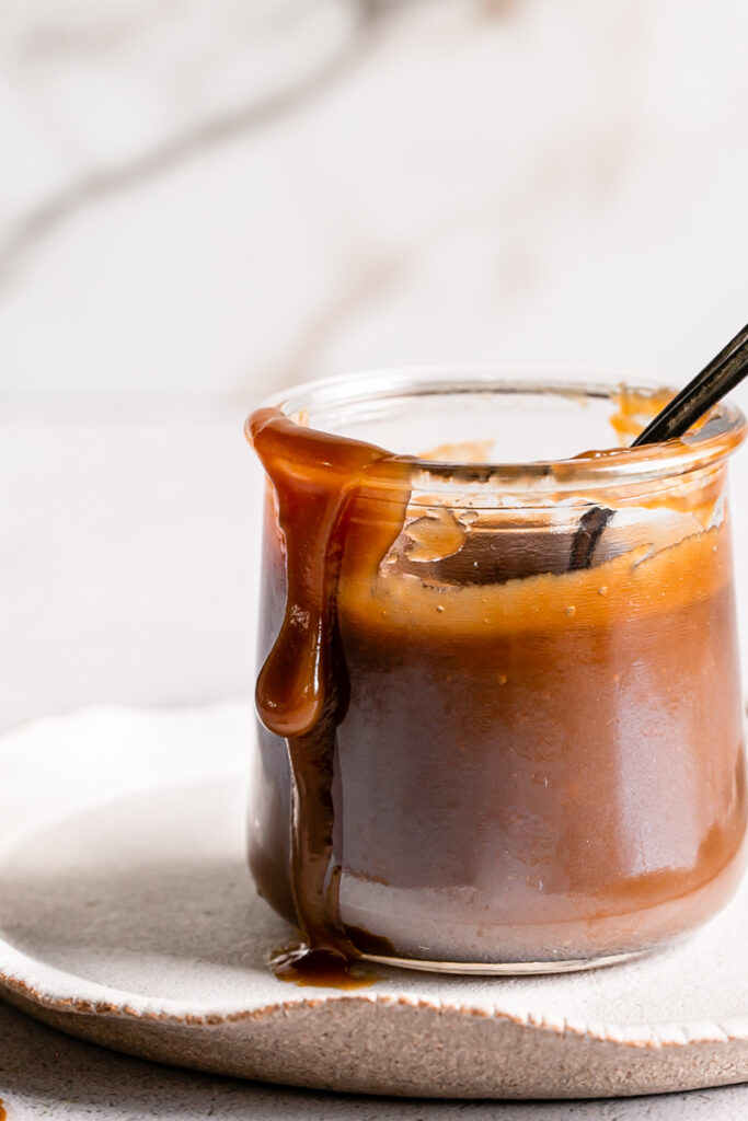 close up of a jar of caramel on a plate with a spoon in it and the caramel dripping down the outside of the glass