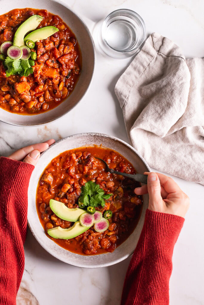 two bowls of vegan three bean harissa pumpkin chili, hands holding one of the bowls spooning some out