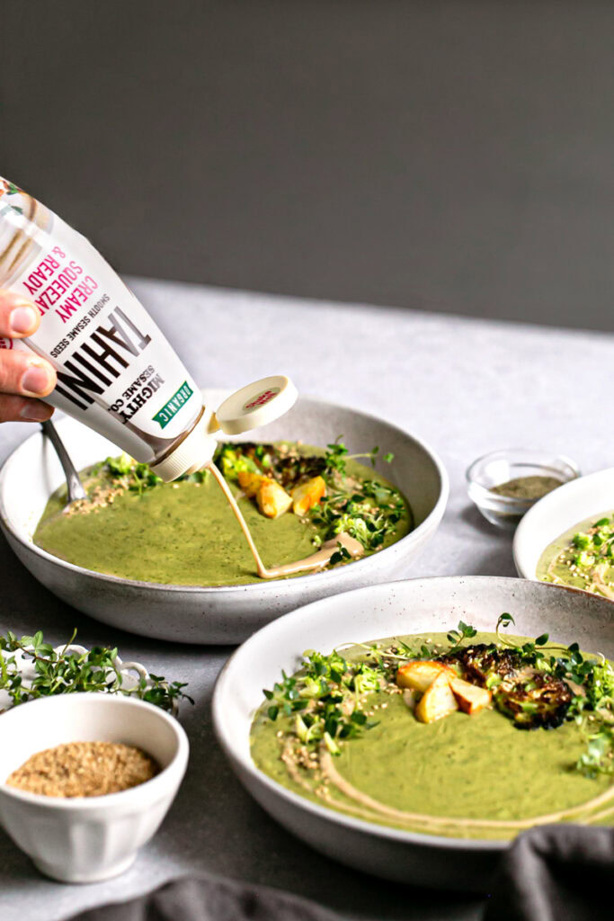 3/4 view of two bowls of roasted broccoli potato tahini soup with squeezing some mighty sesame co tahini right on top