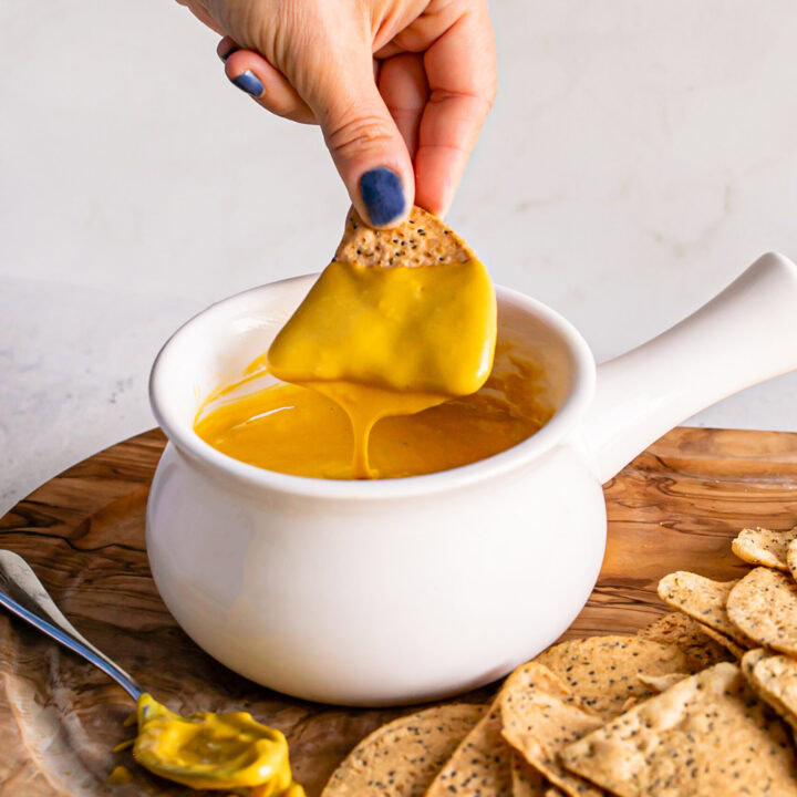 a bowl of vegan nacho cheese sauce with a chip being dipped in it and cheese sauce dripping off of the chip