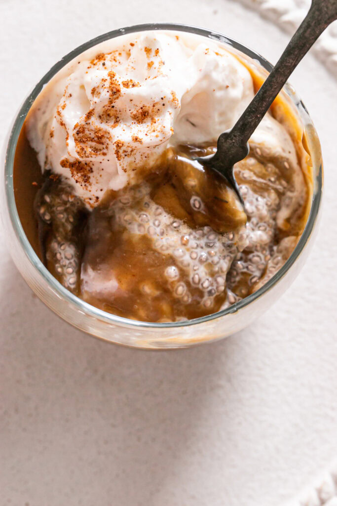 close up overhead shot of of a glass of caramel eggnog chia pudding topped with vegan caramel, maple syrup and whipped cream with a spoonful taken out of it and the spoon in the glass