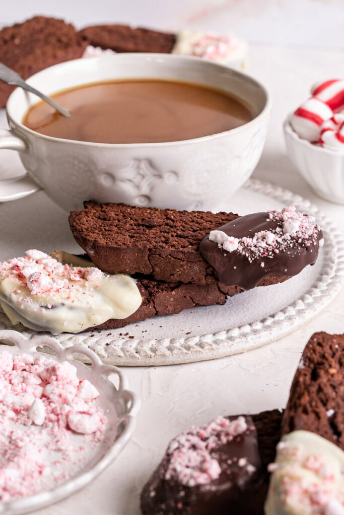 vertical image of Gluten-Free Chocolate Peppermint Biscotti on a plate with a cup of coffee, a plate of crushed peppermints in front of it and a bowl of whole peppermints behind it