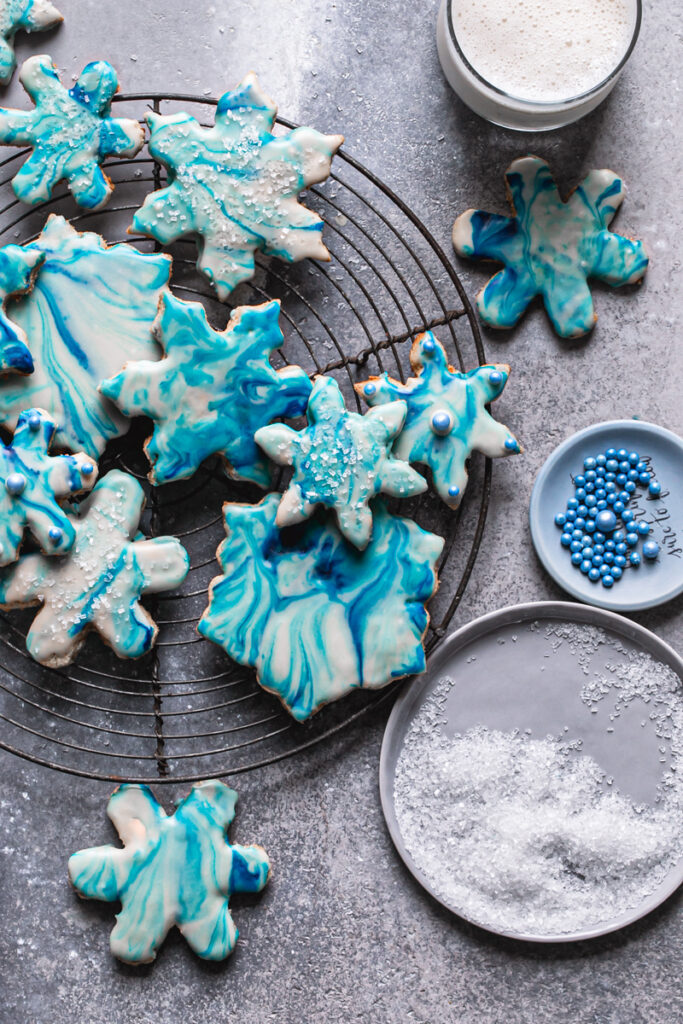 close up of blue and white grain-free marbled snowflake sugar cookies on a round wire cooling rack with two glasses of milk a plate of sparking sugar and blue candy pearls