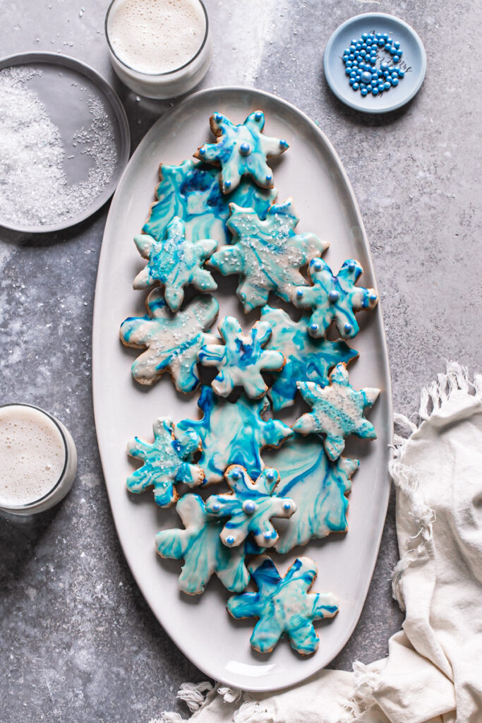 blue and white grain-free marbled snowflake sugar cookies on a white oval plate with two glasses of milk, sparkling sugar, candy pearls and a cloth napkin