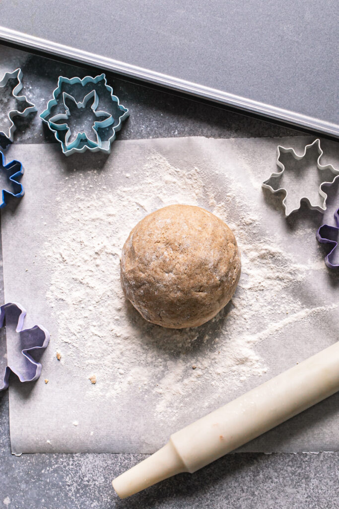 a ball of sugar cookie dough on a piece of parchment paper with a rolling pin, snowflake cookie cutters, and a baking sheet