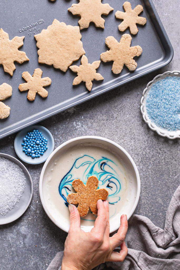 action shot of dipping a snowflake cookie in the blue swirled icing with the un-iced cookies on a baking sheet above it, sparking sugar and candy pearls beside it