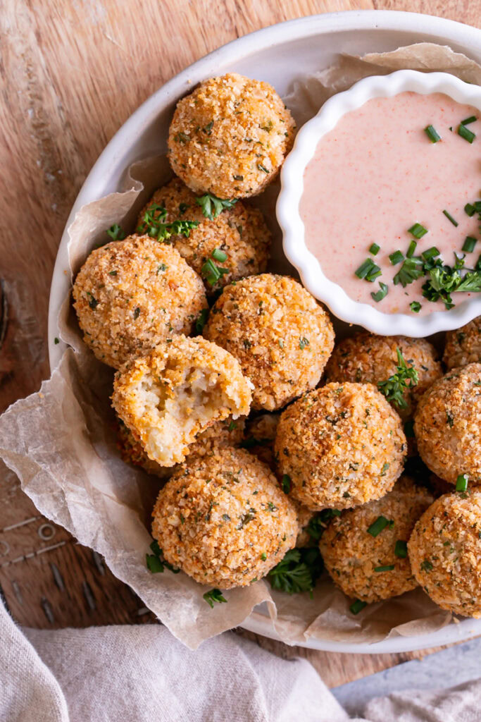 close up image of a bowl full of mashed potato croquettes, one with a bite taken out of it and a bowl of paprika dipping sauce garnished with fresh chives and parsley
