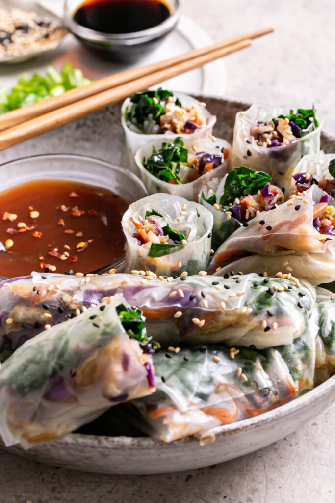 a close up 3/4 view of a bowl of sesame kale spring rolls, some cut in half to reveal the filling, served with a bowl of sweet and sour sauce