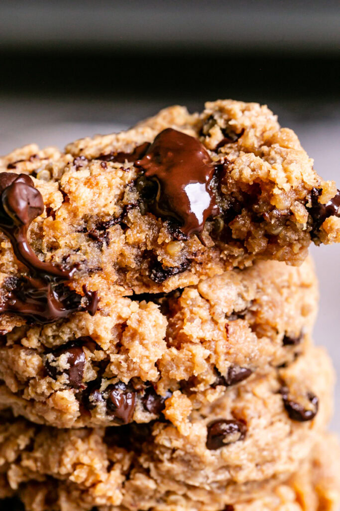 a close up of Quinoa Chocolate Chip Cookies stacked on top of one another, a bite taken out of the top one and melted chocolate oozing out