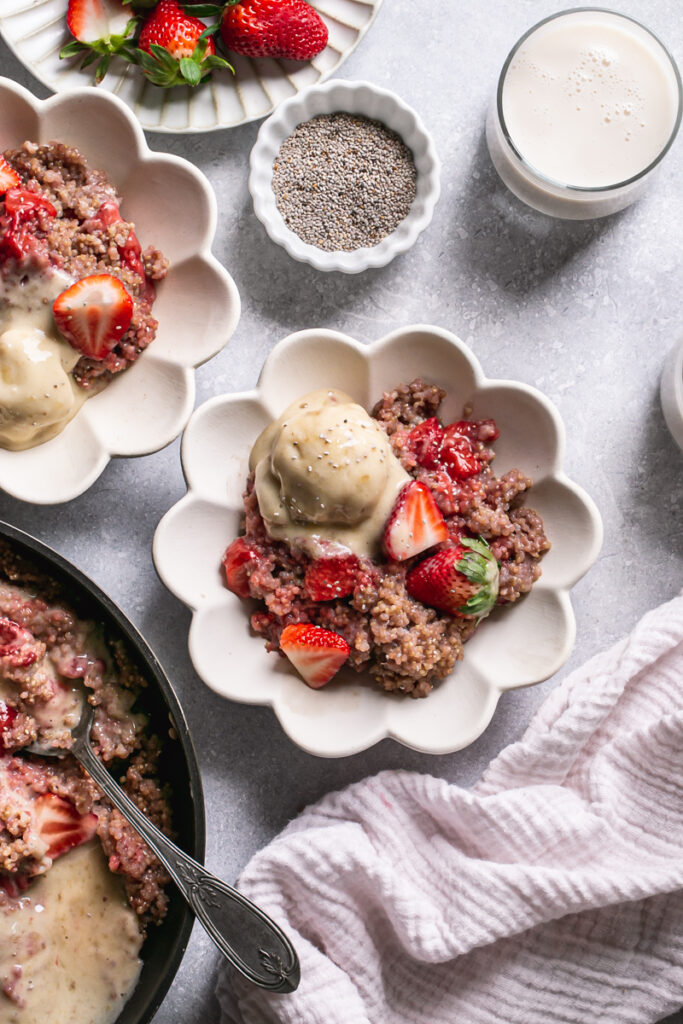 two bowls of strawberries and cream breakfast quinoa with the skillet below it, a plate of strawberries, a glass of almond milk and a small bowl of chia seeds
