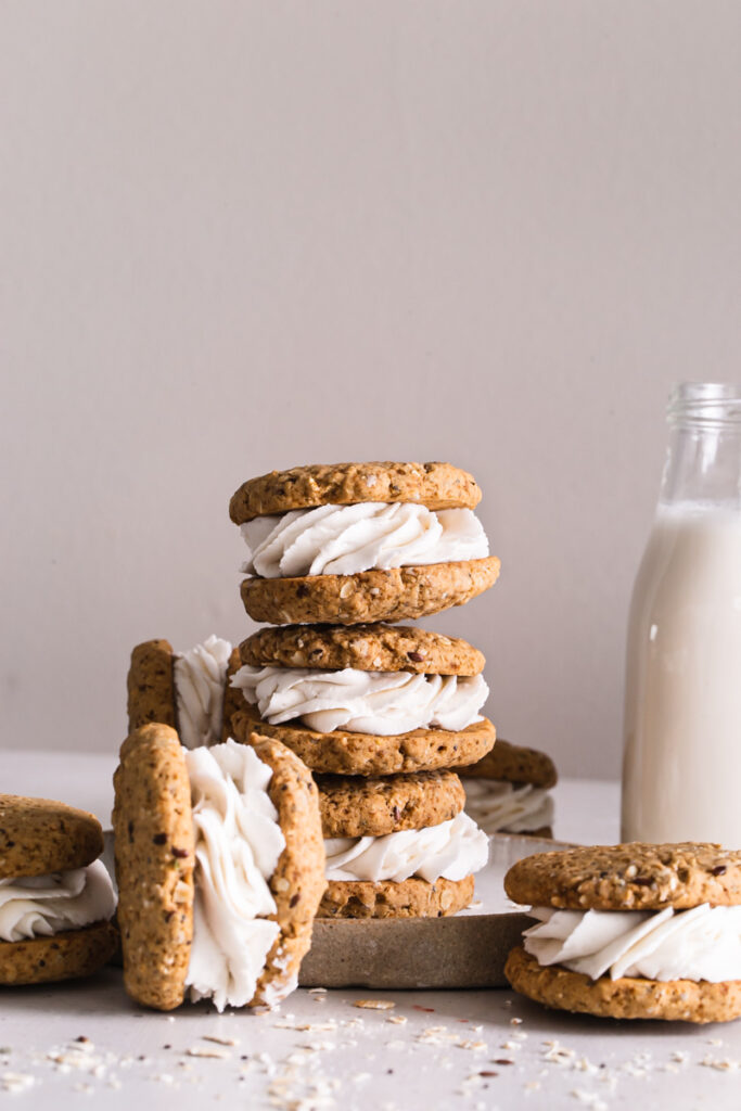 three oatmeal cookie sandwiches stacked on top of one another with another on the side and one in front with a jug of plant milk in the background