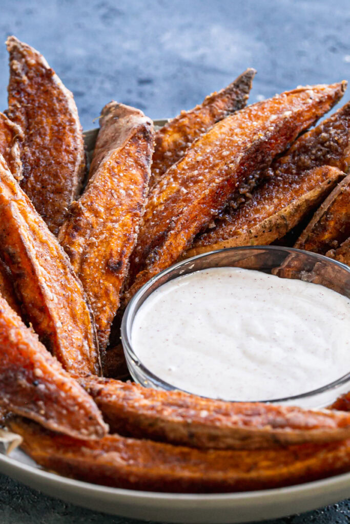 3/4 shot of crispy cinnamon sugar sweet potato wedges in a parchment lined bowl surrounding a bowl of coconut sugar sour cream dipping sauce