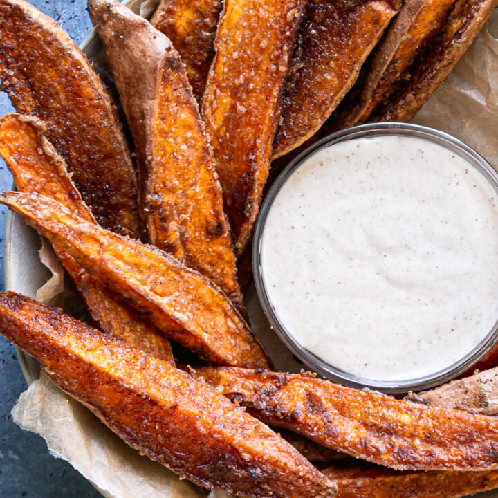 close up shot of crispy cinnamon sugar sweet potato wedges in a parchment lined bowl surrounding a bowl of coconut sugar sour cream dipping sauce