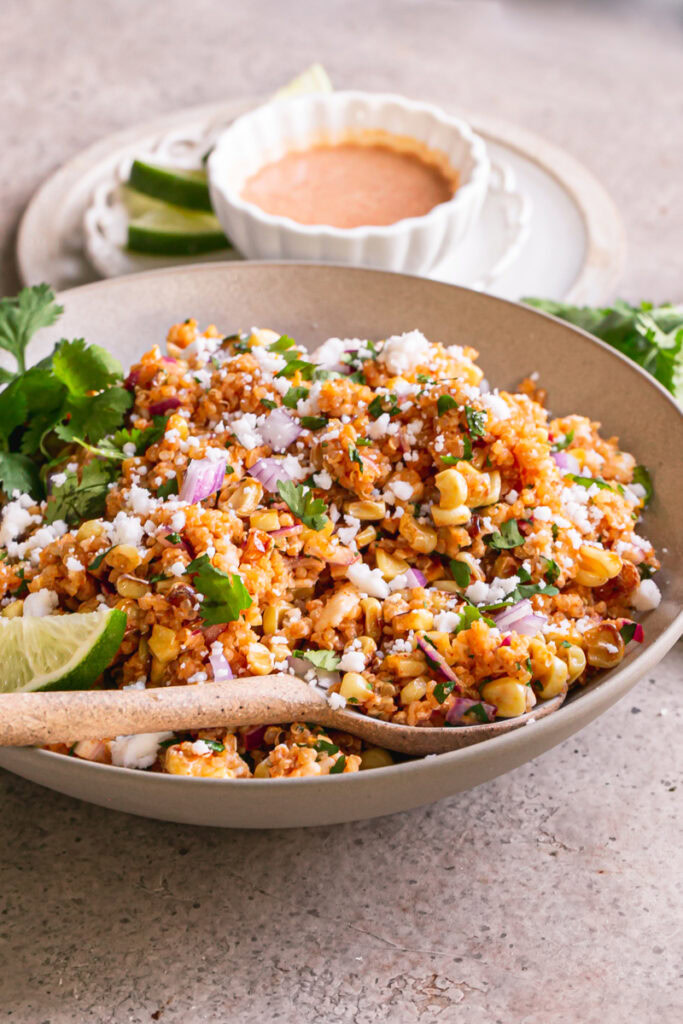 3/4 view of a a bowl of Dairy-Free Mexican Street Corn Quinoa Salad with a spoon in the bowl and a bowl of dressing behind it