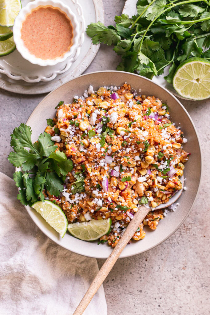 a bowl of Dairy-Free Mexican Street Corn Quinoa Salad with a plate of fresh cilantro leaves and a half of lime above it and a bowl of dressing on the other side