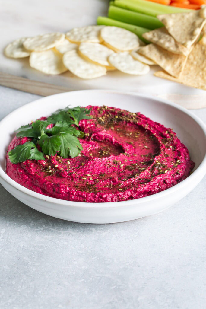 3/4 shot of a bowl of Za'atar Roasted Beet White Bean Hummus garnished with fresh cilantro leaves with rice crackers and tortilla chips on a cutting board in the background