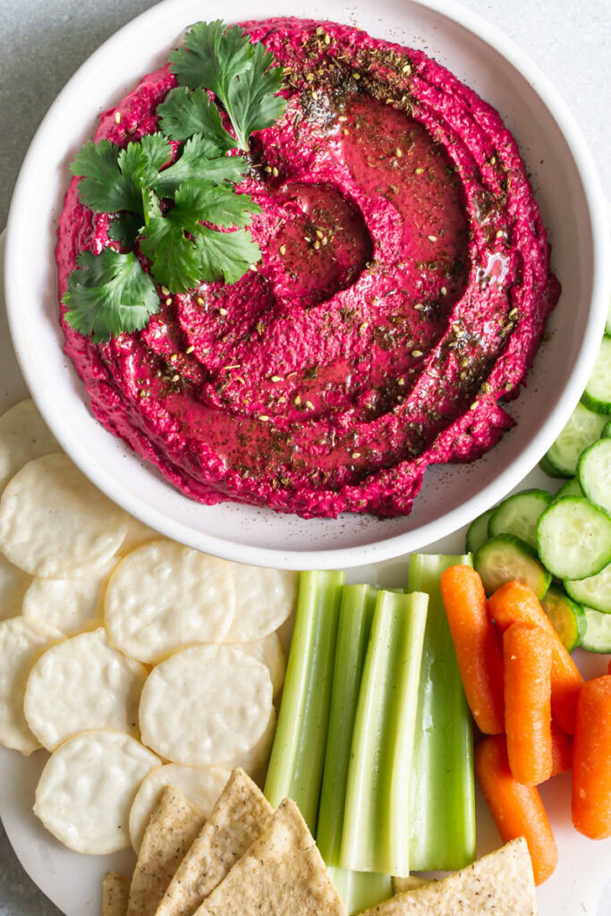 a bowl of Za'atar Roasted Beet White Bean Hummus garnished with fresh cilantro leaves with crackers, chips, celery, carrots, and cucumber around the bowl