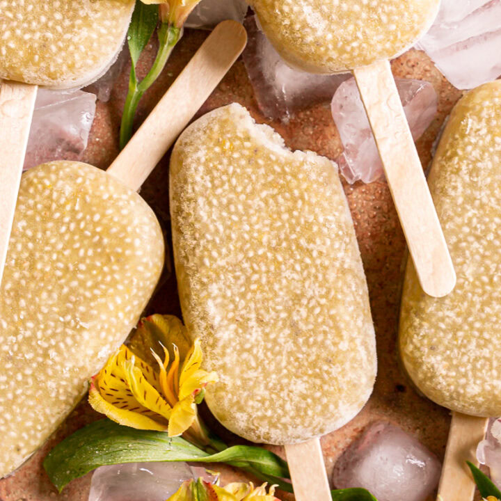 a close up of Pineapple Coconut Chia Popsicles on an orange backdrop covered in ice cubes with yellow and white flowers around them, one popsicle with a bite taken out of it