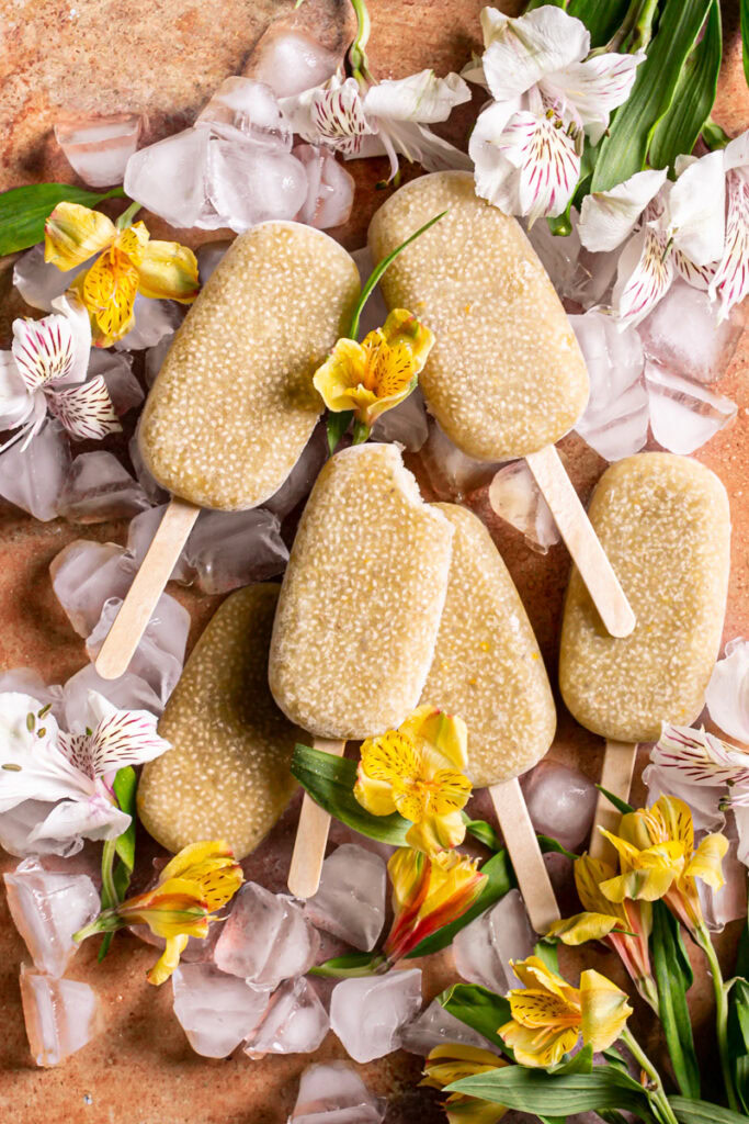 Pineapple Coconut Chia Popsicles on an orange backdrop covered in ice cubes with yellow and white flowers around them