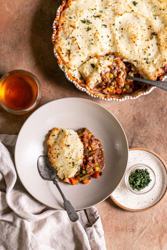 over head shot of Vegan Gluten-Free Shepherd's Pie in a pie baking dish with some spooned into a bowl with a glass of wine, a linen napkin and a bowl of chopped parsley