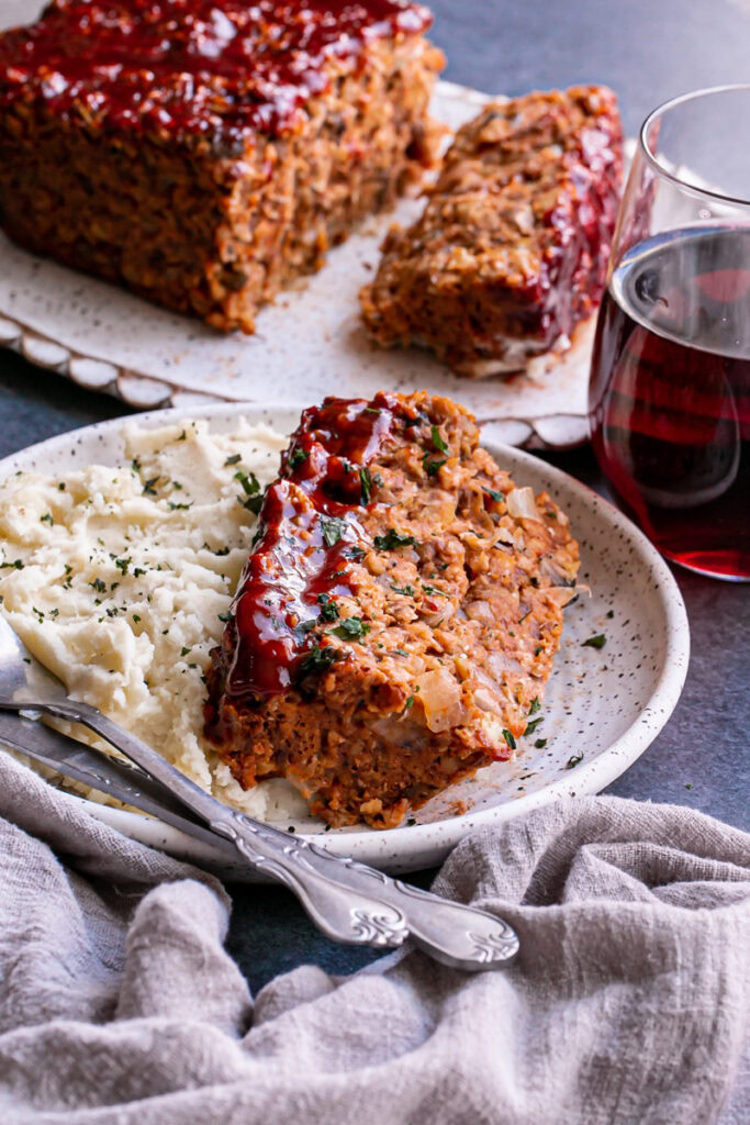 a slice of Vegan Gluten-Free Meatloaf on a plate with mashed potatoes, a glass of red wine and the remaining meatloaf on a platter in the background