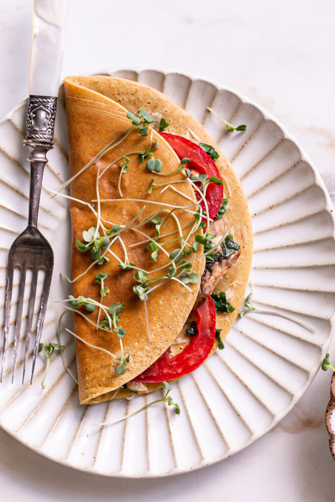A Vegan Gluten-Free Savory Pancake filled with vegan cheese, kale, mushrooms, onion, and tomato on a plate garnished with micro greens