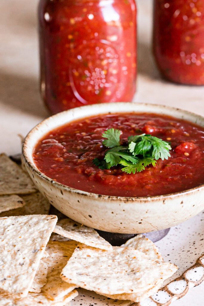 a bowl of salsa garnished with fresh cilantro surrounded by tortilla chips and jars of salsa in the background
