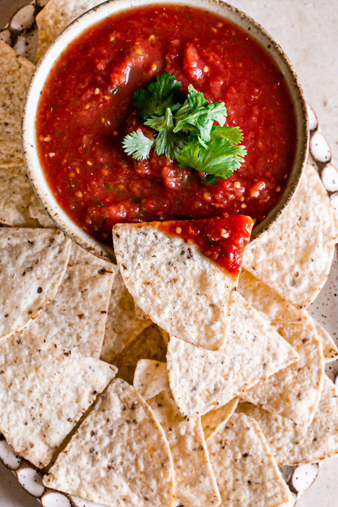 an overhead image of a bowl of salsa garnished with fresh cilantro surrounded by tortilla chip and one chip laying on the rim of the bowl with salsa on it