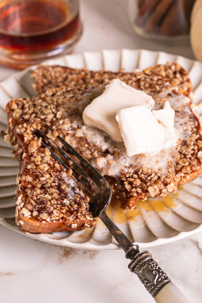 close up of a slice of pumpkin spice oat crusted French toast on a plate with two squares of butter and a fork cutting a slice off